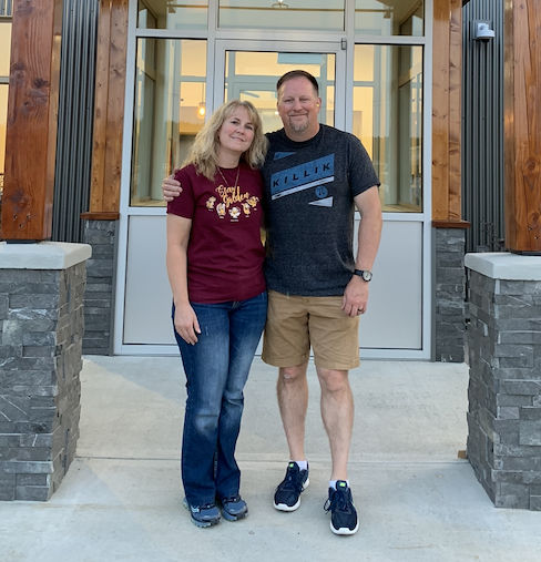 Jim and Debbie Delker smile for the camera under the entryway to their new clinic, which is made of grey brick and wooden pillars. Jim has his left arm around Debbie.