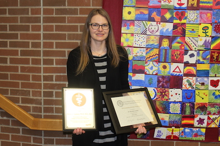 Roxanne Larsen stands smiling at the camera and holding the Zoetis Distinguished Veterinary Teaching Award and the VBS departmental teaching award in front of the quilt made by the Class of 2022.