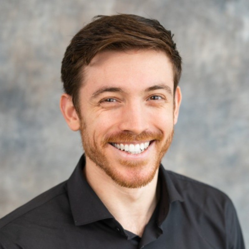 Smiling man with short brown hair and a light beard, wearing a black collared shirt against a textured grey backdrop.