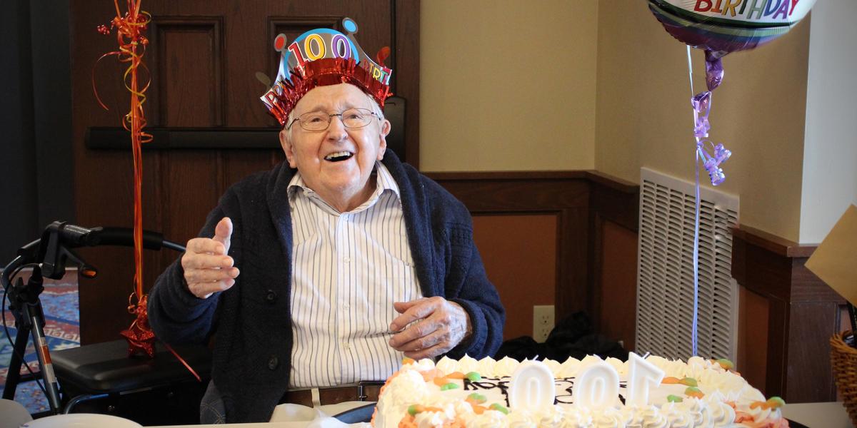 Al Weber smiles at the camera wearing a birthday crown and giving a thumb's up. He is sitting at a table with a white sheet cake on top of it that has "100" shaped birthday candles on it. Next to him, a balloon that reads: "100 BIRTHDAY"