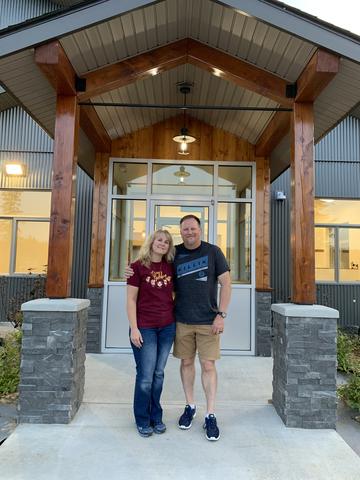 Debbie (left) and Jim (right) smile in front of their new facility. Jim has his left arm around Debbie. 