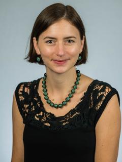 Eva Furrow sits for her faculty portrait. She is wearing a black blouse, a jade necklace, and matching earrings. She has short brown hair and brown eyes. 